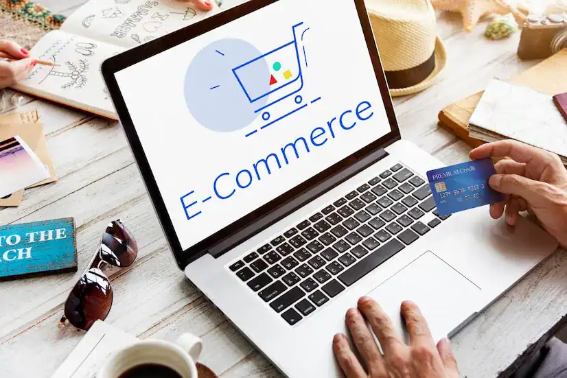 Laptop displaying "E-Commerce" logo with shopping cart, person holding credit card, surrounded by notebooks and sunglasses, emphasizing online retail and financial transactions in e-commerce.