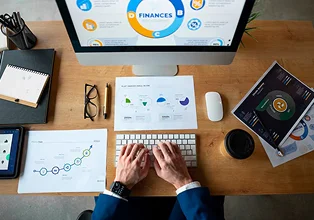 Person typing on a keyboard in front of a computer displaying financial data and charts, with documents and a coffee cup on a desk, illustrating finance optimization for social enterprises.